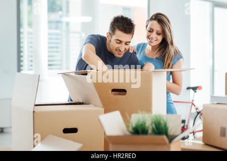 Smiling young couple unpacking carton boxes in a new house, he is searching objects in a big box on a table Stock Photo