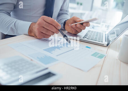 Businessman at desk using a financial app on his smart phone and working on reports, hands close up Stock Photo