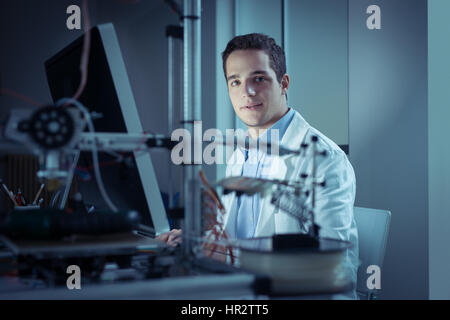 Young researcher in the lab, wearing a lab coat and working with a computer, 3D printer on foreground, he is looking at camera Stock Photo