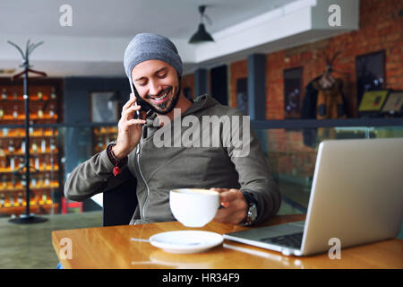 Young handsome hipster man with beard sitting in cafe talking mobile phone, holding cup of coffee and smiling. Laptop on wooden table. Stock Photo