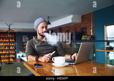Young handsome hipster man with beard sitting in cafe with a cup of coffee, vaping and releases a cloud of vapor. Working at laptop and having a little break. With copy space. Stock Photo