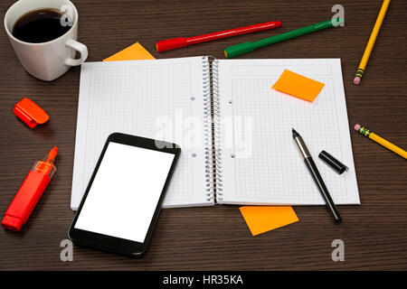 Opened notebook, smartphone with blank screen, pen, sticky note, cup of coffee in chaos on dark office wooden desk. Stock Photo