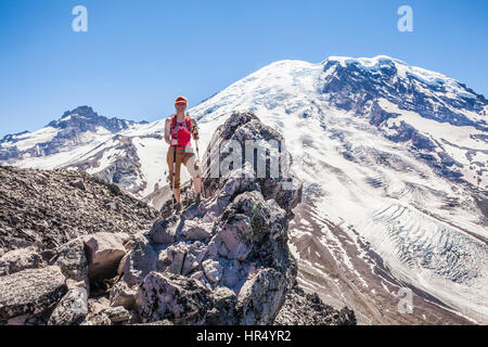 A woman atop a rocky outcrop on Burroughs Mountain with Mount Rainier looming above in the background.  Rainier National Park, Washington, USA. Stock Photo