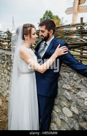 Bride hugs groom near stone wall with wooden fence at wedding day. Stock Photo