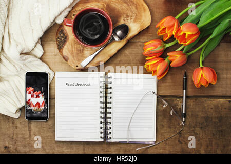 Overhead shot of an open food journal book with cell phone, coffee and flowers over a wood table top ready to plan diet. Flat lay top view style. Stock Photo