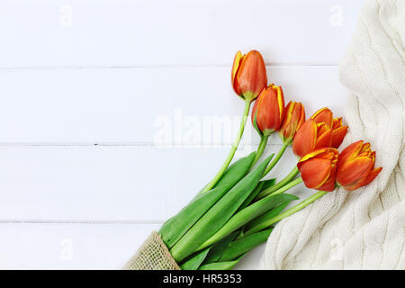Overhead shot a bouquet of orange and yellow tulips wrapped in toile and burlap fabric and a cozy knit throw blanket over white wood table top. Flat l Stock Photo