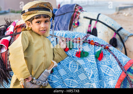 Muscat, Oman - Feb 4, 2017: Young Omani boy dressed in traditional clothing posing next to his camel. Stock Photo