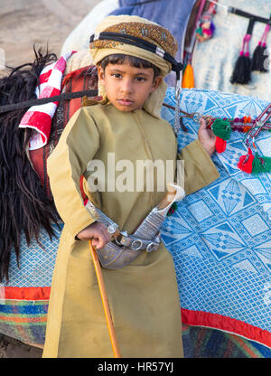 Muscat, Oman - Feb 4, 2017: Young Omani boy dressed in traditional clothing posing next to his camel. Stock Photo