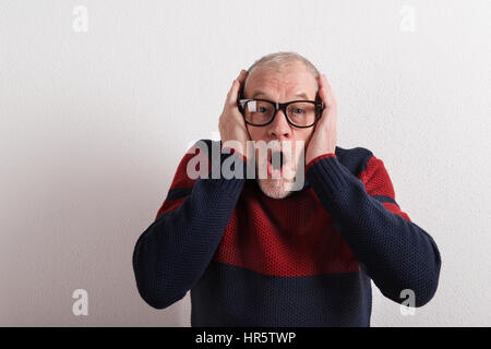 Shocked senior man in red and blue sweater and black eyeglasses holding head. Studio shot against white wall. Stock Photo