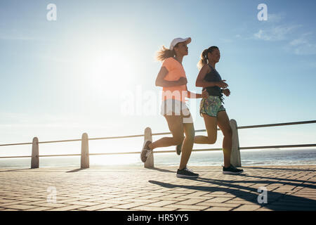 Low angle shot of two young women running on ocean front. Female runner working out on a sunny day. Stock Photo