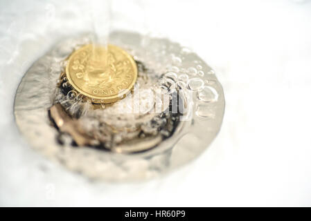 British money including a one pound coin stuck in a sink drain plughole as water flows over the coins. Stock Photo