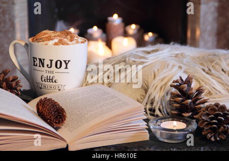 Reading by candlelight by an open fireplace in a cosy English home with a mug of creamy coffee and a blanket (throw) on a winter evening Stock Photo