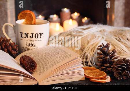 Reading a paperback book by an open fire in a cosy hygge inspired English living room with a hot drink and blanket (throw) on an autumn night Stock Photo