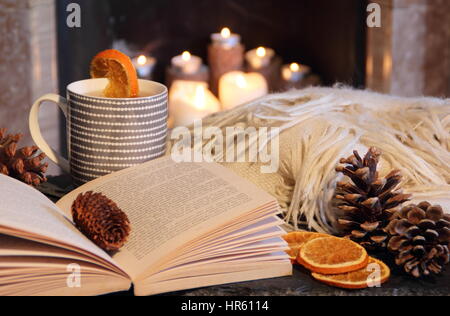 Reading a paperback book by a fireplace in a cosy hygge inspired English home with a mug of hot tea and a blanket (throw) in autumn Stock Photo