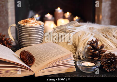 Reading a paperback book by candle light next to an open fireplace in a cosy English home with a creamy hot drink and blanket on a winter evening Stock Photo