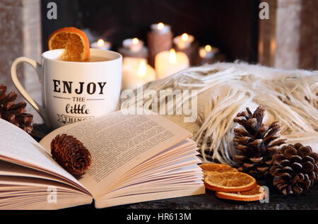 Reading a paperback book by an open fireplace in a cosy hygge inspired English living room with a hot drink and blanket (throw) - autumn/winter Stock Photo
