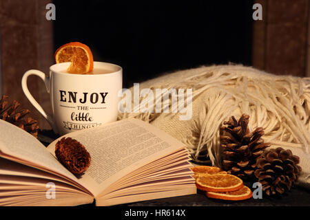 Reading a paperback book by a fireplace in a cosy hygge inspired English living room with a hot drink and blanket (throw) - autumn/winter Stock Photo