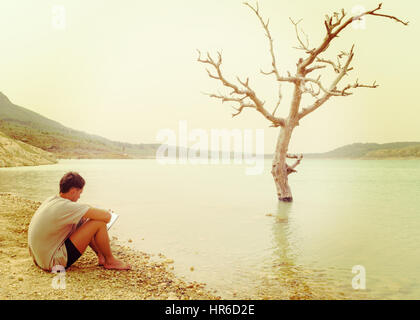 good looking male hipster sat by tranquul lake reading a book in shorts and tshirt on a hot summer day.retro filter, conceptual dead tree in the lake. Stock Photo