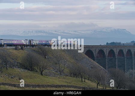 An Arriva Rail Northern passenger diesel train going across the Ribblehead Viaduct on a grey winter day with snow-capped Pen-y-Ghent beyond.