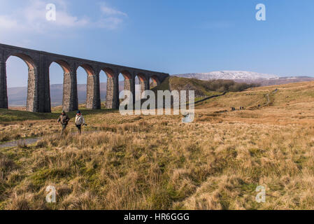 Under blue sky, couple walk on footpath by towering Ribblehead Viaduct on sunny winter day (snow covered Whernside beyond) - Yorkshire Dales, GB, UK. Stock Photo