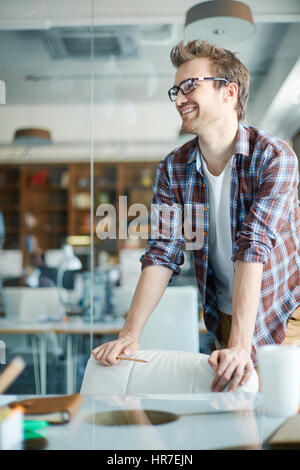 Young stylish man wearing checkered shirt and glasses standing leaning on chair in modern airy office space, talking to his colleagues and smiling Stock Photo