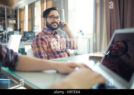Successful Asian businessman dressed in casual checkered shirt, sitting back in chair in light spacious office and talking on phone, while his colleag Stock Photo