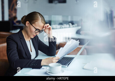 Young female executive busy reading documents focused on her work looking troubled and thinking hard seated in lounge area of modern restaurant at cof Stock Photo