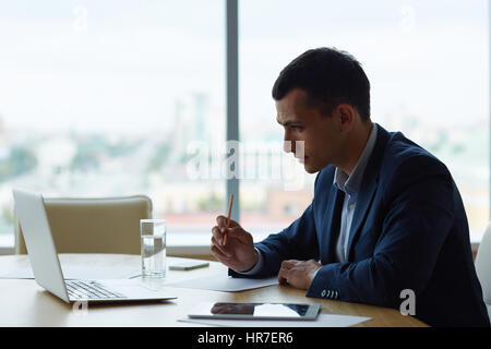 Profile view of young business man sitting against window at table in office, working with laptop and making notes on paper looking focused Stock Photo