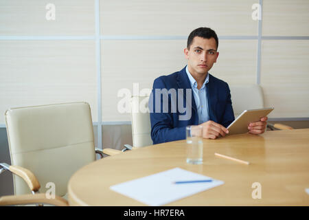 Young serious business man sitting alone at large table in office, looking up to camera from working with tablet while preparing for upcoming meeting Stock Photo