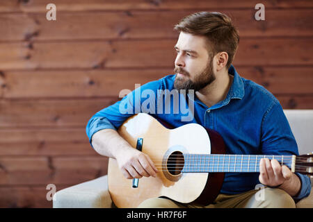 Portrait of young bearded man in denim shirt playing acoustic guitar and looking away thoughtfully Stock Photo