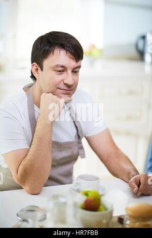 Portrait of middle-aged man in apron sitting at dinner table and looking away dreamily with smile Stock Photo