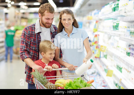Pretty young woman in blue polo T-shirt posing for camera with wide smile and holding milk bottle in hand while her family looking away Stock Photo