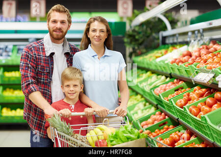 Cheerful family of three with full shopping trolley posing for camera in fruit and vegetable department of supermarket Stock Photo