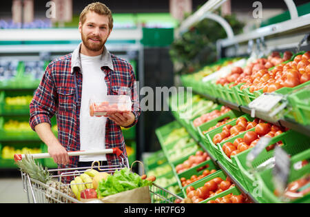 Young bearded bachelor holding packaging box with cherry tomatoes and looking at camera while shopping in fruit and vegetable department of hypermarke Stock Photo