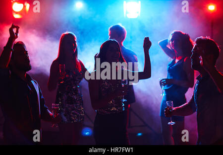 Silhouettes of young dressed-up people dancing in club smoke with glasses of champagne Stock Photo