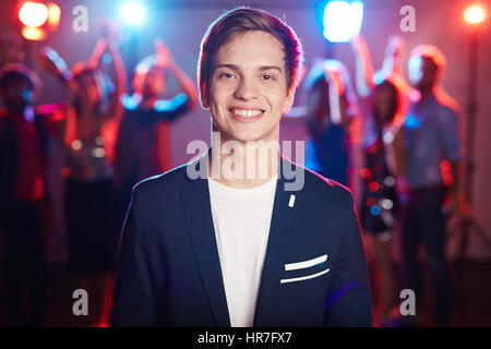 Portrait of confident blond young man in jacket looking at camera with nice toothy smile against dancing crowd background Stock Photo