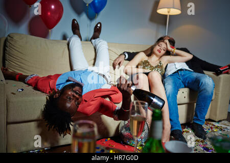 Group of glamorous friends relaxing on sofa after wild house party, they holding champagne and beer bottles in hands while having nap Stock Photo