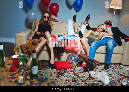 Messy room after wild house party, three tipsy stylish friends relaxing on couch while blond-haired woman with teddy bear posing for photography Stock Photo
