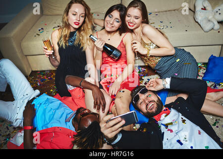 Middle-aged Asian man in sunglasses lying on floor covered with confetti and taking selfie with his beautiful female friends Stock Photo