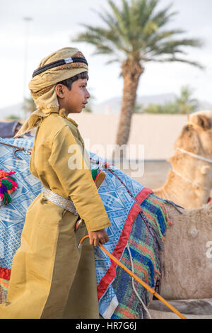 Muscat, Oman - Feb 4, 2017: Young Omani boy dressed in traditional clothing posing next to his camel. Stock Photo