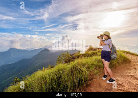 Hiker asian teen girl wear cap and eyeglass with happy holding dslr camera shooting photo beautiful nature in winter on peak mountain at sunset Stock Photo