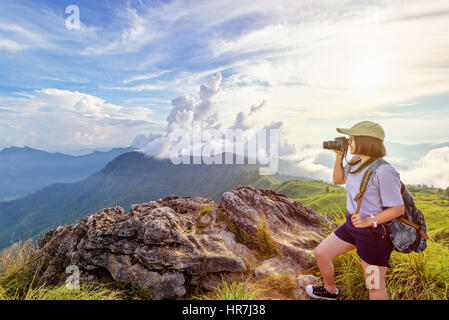 Hiker asian teen girl wear cap and eyeglass with happy holding dslr camera shooting photo beautiful nature in winter on peak mountain at sunset Stock Photo