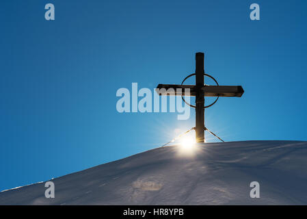 Wooden cross on snow capped mountain hill, setting sun beaming through Stock Photo