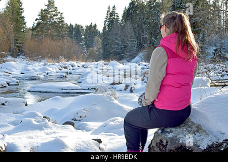 Young woman sitting on stone and watching beautiful small river on winter. Stock Photo