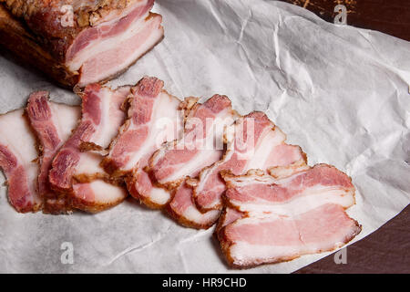 Close up view slices of smoked bacon on the white packaging paper. Naturmort in a rustic style on brown vintage background. Stock Photo