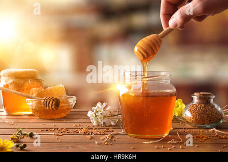 Hand with dipper picking honey from a jar of honey. Jars of honey, bee honeycomb and bee pollen on wooden table with flowers in a rustic kitchen and s Stock Photo