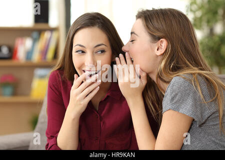 Portrait of a gossip girl telling a secret in the ear to her friend sitting on a couch at home Stock Photo