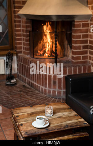 Coffee cup on a table with an open fireplace burning wood in the background Stock Photo