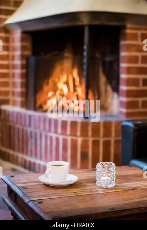Coffee cup on a table with an open fireplace burning wood in the background Stock Photo