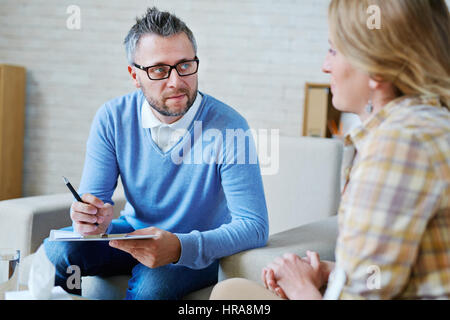 Portrait of middle-aged psychologist in eyeglasses holding clipboard and pen and listening attentively to his female patient Stock Photo